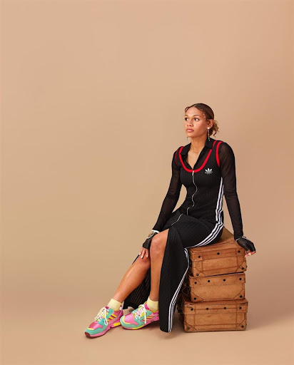Adult female sitting on a stack of suitcases in a black Adidas track suit dress and colorful sneakers in a studio setting.