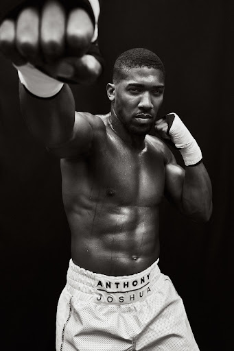 A shirtless Anthony Joshua with white boxing wraps raises his fists in a fighting stance against a stark black background in a studio shot.