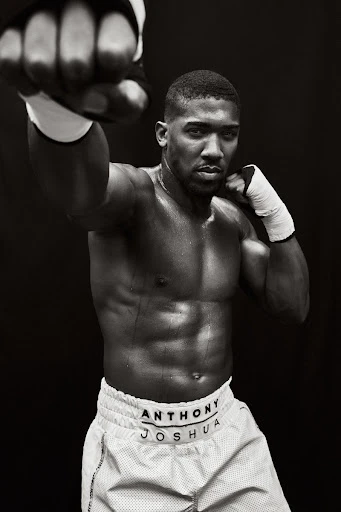 A shirtless Anthony Joshua with white boxing wraps raises his fists in a fighting stance against a stark black background in a studio shot.