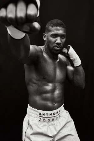 A shirtless Anthony Joshua with white boxing wraps raises his fists in a fighting stance against a stark black background in a studio shot.