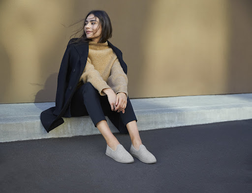Brunette woman in tan sweater and black coat seated against a wall, wearing light grey casual shoes and dark pants, looking left.