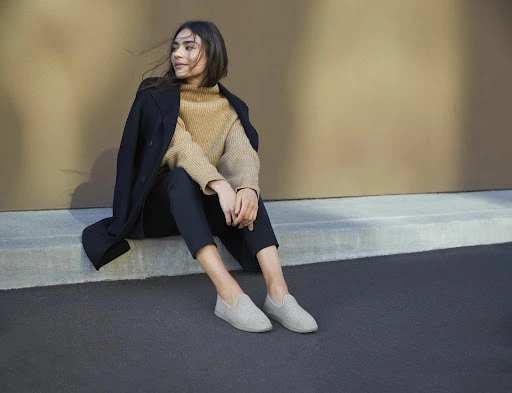 Brunette woman in tan sweater and black coat seated against a wall, wearing light grey casual shoes and dark pants, looking left.