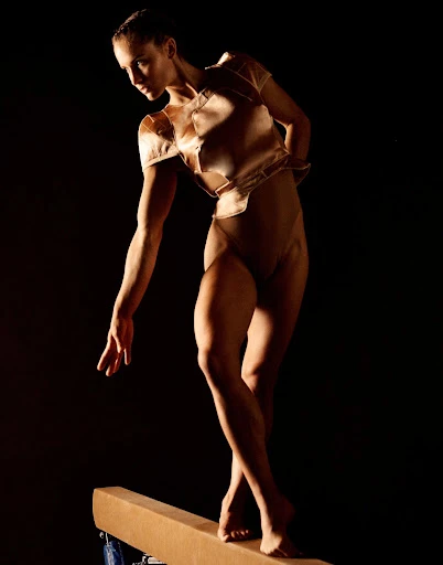 A woman in sportswear balances on a balance beam with a dark background, illuminated by a spotlight.
