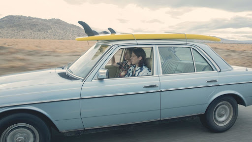 Two people ride in a vintage blue car with surfboards strapped on top, cruising down a desert road with mountains in the distance.