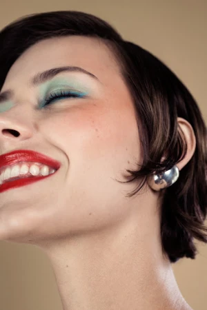 Close-up portrait of a woman with red lipstick, blue eyeshadow, and a silver earring against a neutral background.