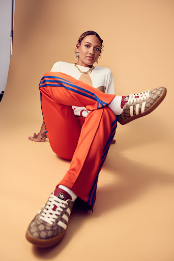 Stylish woman in trendy sportswear poses in a studio, showcasing orange track pants and patterned Adidas sneakers, fashion forward and modern.