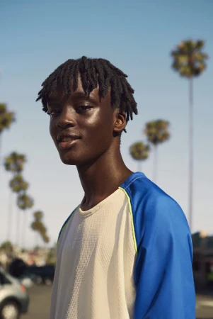 Young Black man with short locs wearing a blue and white raglan t-shirt outdoors against a clear blue sky with distant palm trees