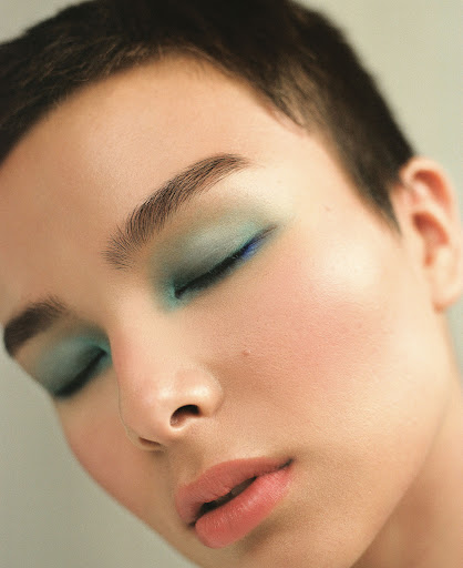 Close-up portrait of a young woman with closed eyes and short dark hair, wearing turquoise eyeshadow and light pink lipstick in studio light.