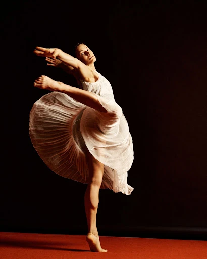 Female dancer in mid-air wearing flowing white dress against a dark backdrop, illuminated by focused studio lighting on red stage.