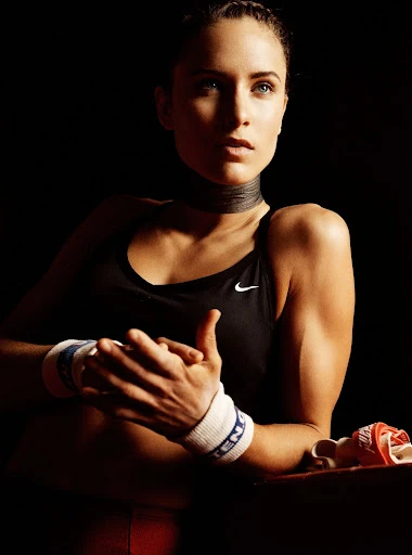 Athletic woman in sports bra and wristbands poses against a dark background, looking off-camera with strong lighting and neutral expression.