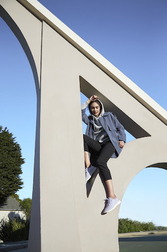 Woman in streetwear poses near a large concrete architectural sculpture against a clear blue sky, showcasing casual fashion.