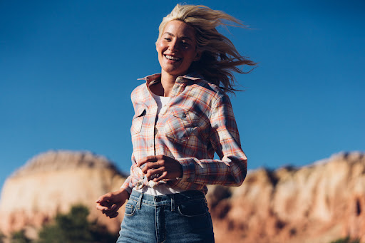 Smiling woman with blonde hair, wearing a plaid shirt and jeans, stands against a backdrop of a clear blue sky and rock formations.