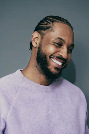 Portrait of a smiling adult male with cornrow braids, a beard, and a light purple sweater against a solid gray background.