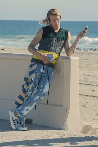 Young man in casual streetwear stands by a beach listening to music on headphones, wearing a knit vest and splattered jeans on a sunny day.