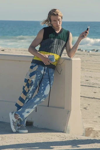 Young man in casual streetwear stands by a beach listening to music on headphones, wearing a knit vest and splattered jeans on a sunny day.