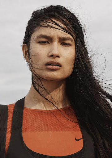 Close-up portrait of a woman with wet dark hair wearing an orange and black athletic top against a pale sky.