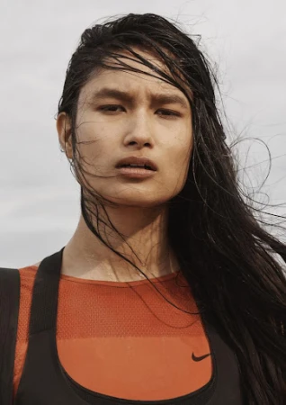 Close-up portrait of a woman with wet dark hair wearing an orange and black athletic top against a pale sky.