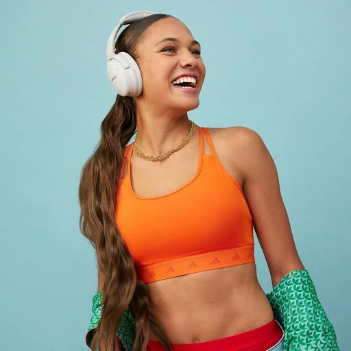 Smiling young woman with long brown hair, wearing headphones and a sports bra, set against a light blue backdrop.