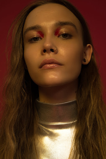 Close-up portrait of a woman with long brown hair, glossy lips, and striking red eyeshadow against a solid red background.
