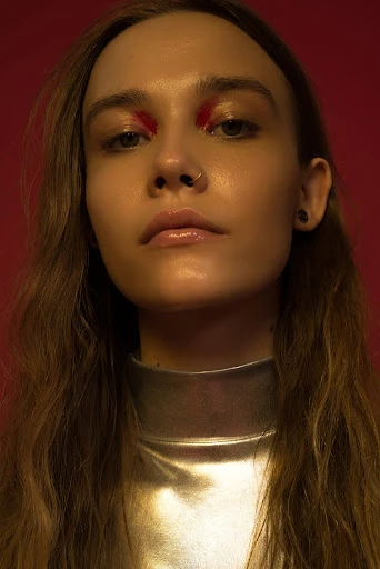 Close-up portrait of a woman with long brown hair, glossy lips, and striking red eyeshadow against a solid red background.
