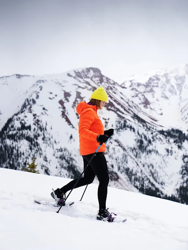 Woman snowshoeing through a winter landscape with snow-covered mountains, wearing a bright orange jacket and yellow beanie.
