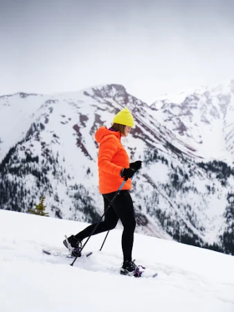Woman snowshoeing through a winter landscape with snow-covered mountains, wearing a bright orange jacket and yellow beanie.