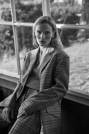 Blonde woman poses in a plaid suit near a window with a contemplative expression in a black and white portrait. Fashion editorial.
