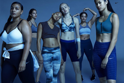 Group of diverse women in sports bras and leggings posing in a studio with Nike logos, promoting fitness and athletic wear.