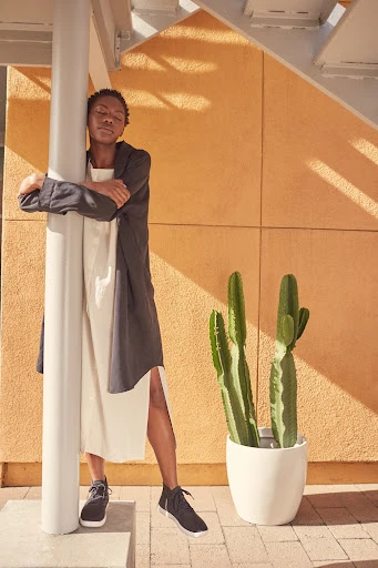Woman in a neutral dress and jacket leans against a white column next to a potted cactus in a sunlit outdoor setting.