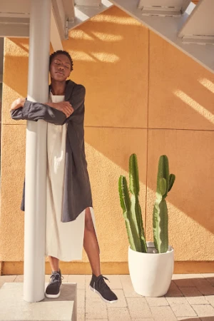 Woman in a neutral dress and jacket leans against a white column next to a potted cactus in a sunlit outdoor setting.