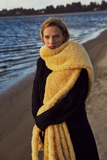 Woman wrapped in a thick yellow scarf and dark sweater standing on a sandy beach near water