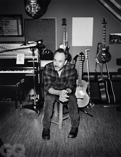 Black and white portrait of a man with a flannel shirt sitting on a stool, surrounded by musical instruments in a music studio.
