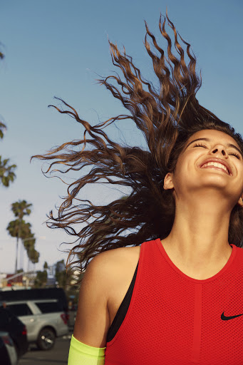 An athletic woman with flowing brown hair smiles in the sun, wearing a red tank top with a black logo and a neon yellow arm sleeve.
