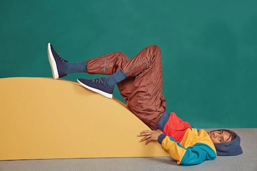Person lounges on a yellow structure in a hooded sweatshirt and trousers wearing navy sneakers against a turquoise background.