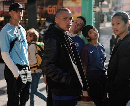 Group of five people in urban setting, wearing casual clothing and posing outdoors. Daylight illuminates the city street backdrop.