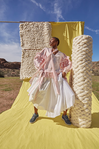 A woman in a pink polka dot top and white skirt poses in a field against a yellow backdrop with wool props on a sunny day.