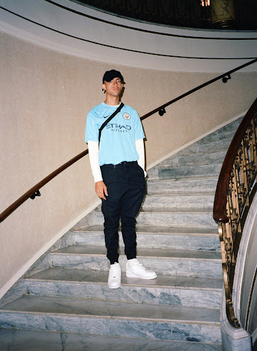 Stylish young man on marble stairs, wearing a light blue soccer jersey, dark trousers, white high-top sneakers, and a black baseball cap.