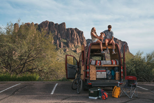 A couple sits on the back of their van in a parking lot with the open doors revealing gear and desert mountains in the background.