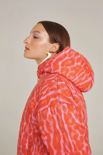 Woman in profile wearing a bright orange and pink patterned hooded puffer coat in a studio setting with neutral background.
