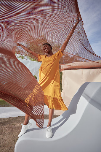 A smiling African-American woman in a yellow dress and white sneakers raises a brown mesh net, posing outdoors on a white structure.
