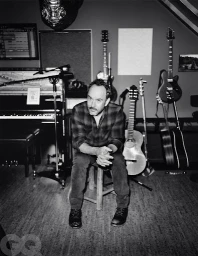 Monochrome portrait of a man seated on a stool inside a music studio surrounded by guitars and a piano.