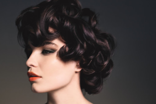 Close-up studio shot of a young adult female model with dark, curly hair, dramatic eye makeup, and orange lipstick on a grey background.