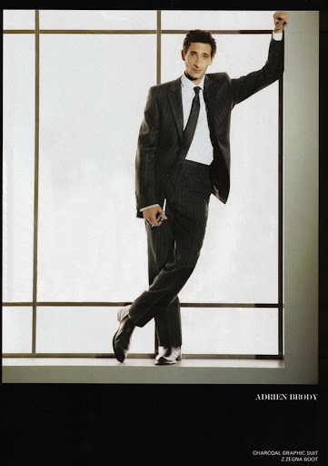 Adrien Brody in a charcoal striped suit leans against a window, looking at the camera. He wears a dark tie and black leather shoes.