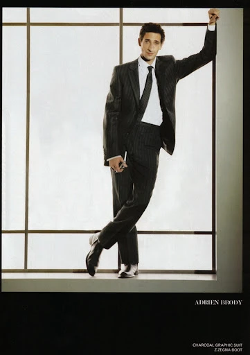 Adrien Brody in a charcoal striped suit leans against a window, looking at the camera. He wears a dark tie and black leather shoes.
