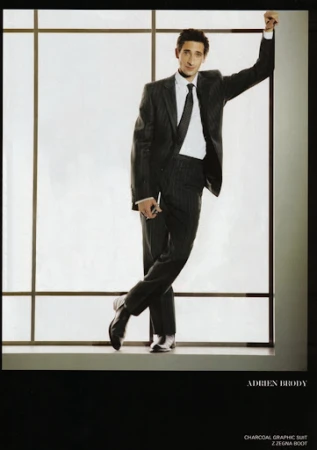 Adrien Brody in a charcoal striped suit leans against a window, looking at the camera. He wears a dark tie and black leather shoes.