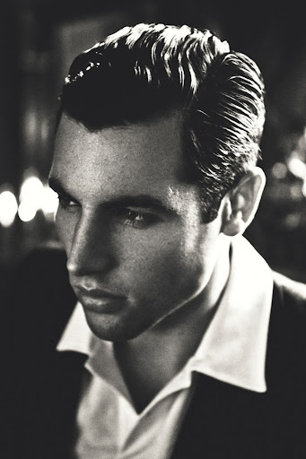 Monochrome portrait of a dark-haired adult male in suit and collared shirt looking off camera with a serious expression in soft studio light.