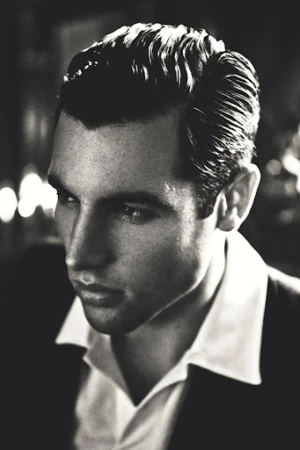 Monochrome portrait of a dark-haired adult male in suit and collared shirt looking off camera with a serious expression in soft studio light.
