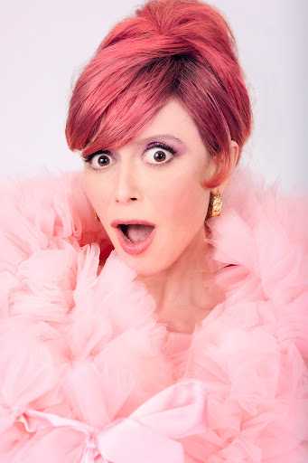 Close-up studio shot of a woman with pink hair wearing a voluminous pink tulle garment, expressing a surprised facial expression.