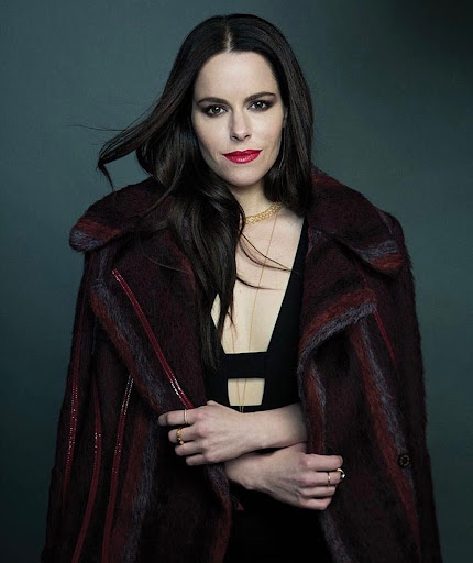 Portrait of a woman with dark hair wearing a burgundy patterned coat over a black top with red lipstick.