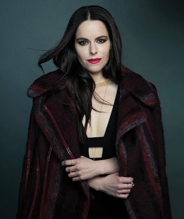 Portrait of a woman with dark hair wearing a burgundy patterned coat over a black top with red lipstick.